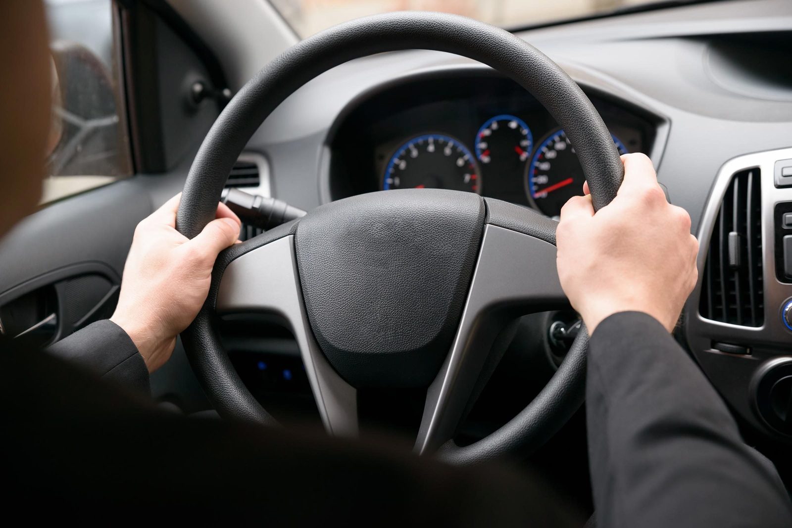 A person grips a steering wheel, driving a car. The interior features a dashboard with various gauges and controls, hinting at a modern vehi