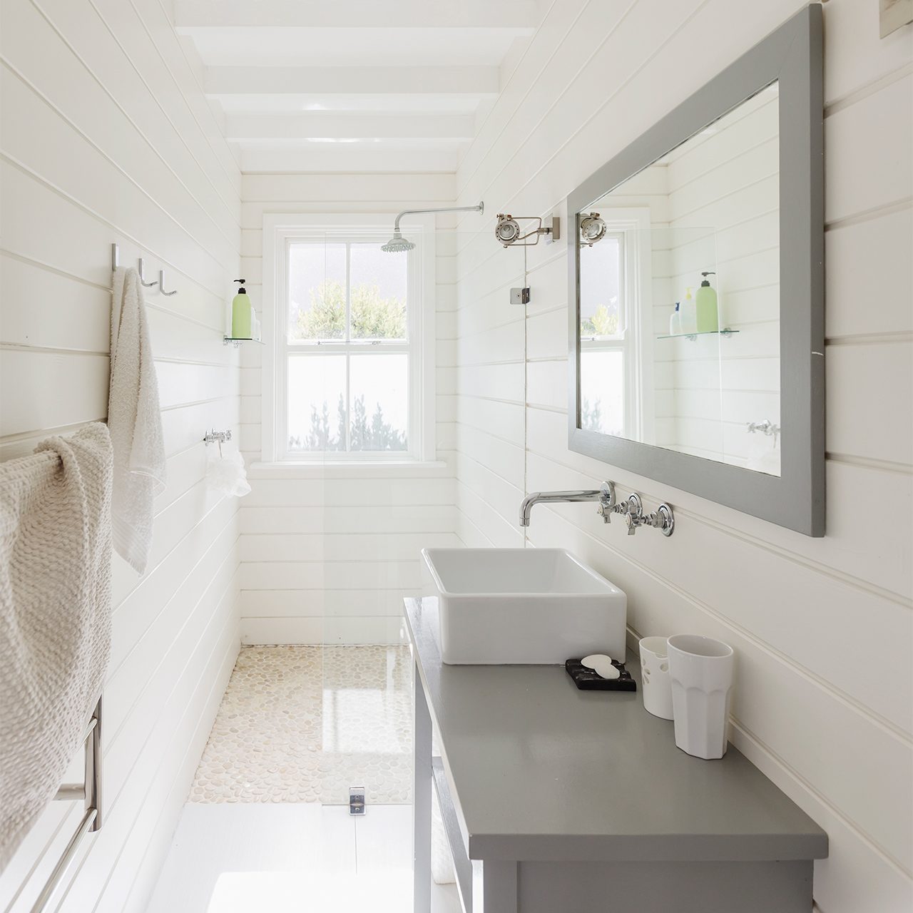 Bright, minimal bathroom with white shiplap walls, large mirror, gray vanity, vessel sink, towel, and two green bottles by a window letting in natural light.