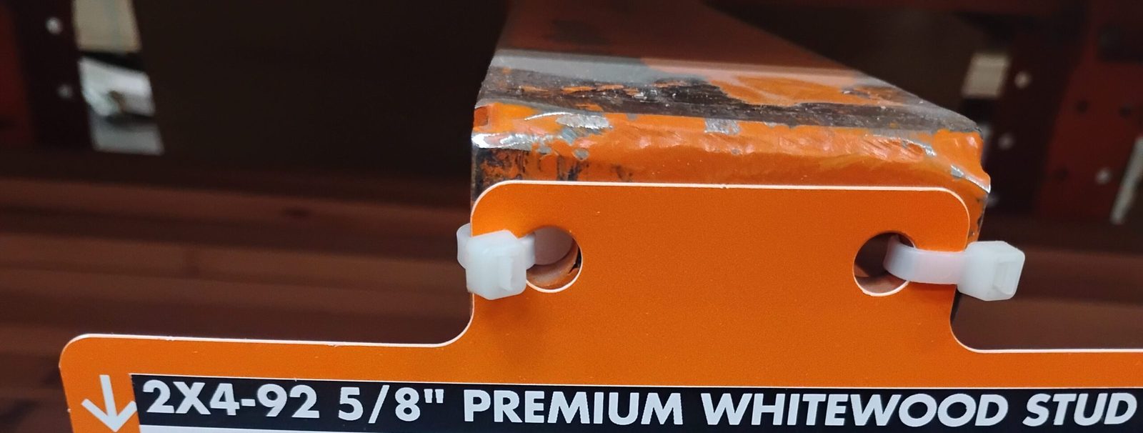 An orange store shelf tag reading 2 by 4, Premium Whitewood Stud, zip-tied to a lumber rack beam.