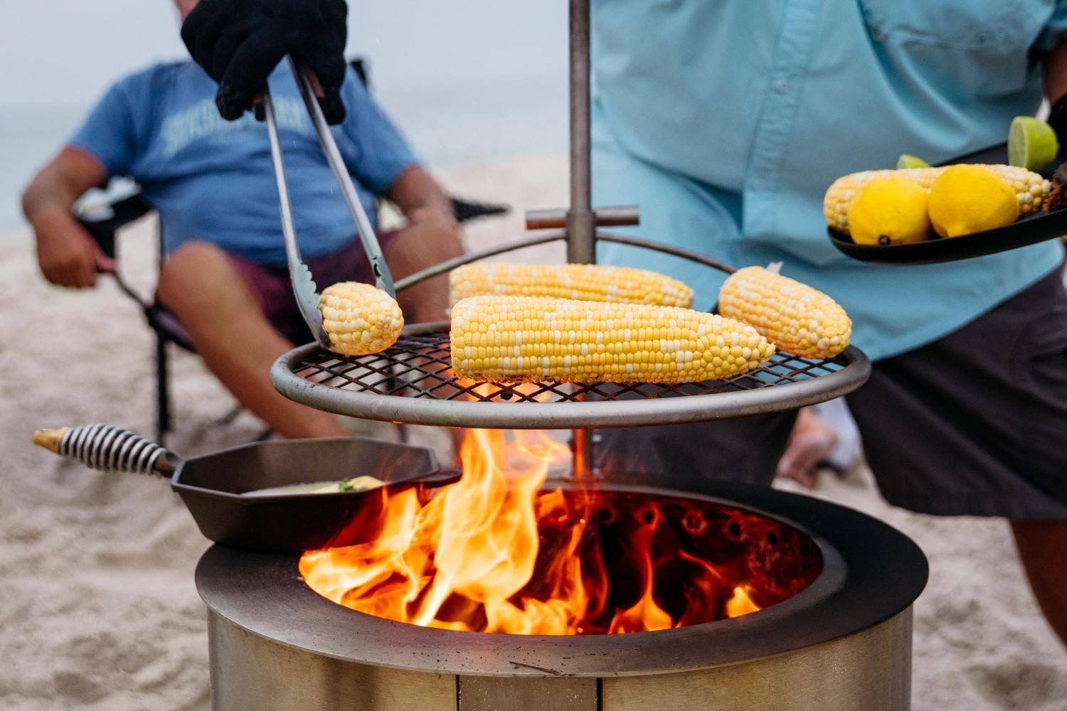 Corn on the grill is being turned over tongs above roaring flames, with a person relaxing nearby on a sandy beach.