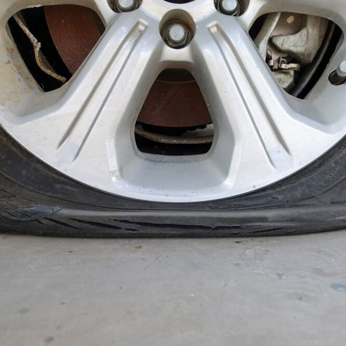 A silver car wheel with a flat tire sits on a flat surface, showing a damaged sidewall and visible brake components behind it.