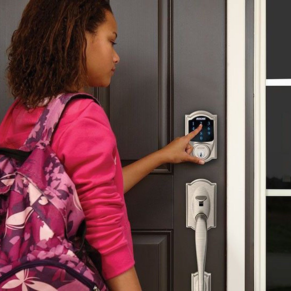 A girl in a pink shirt presses a keypad on a smart door lock while carrying a pink backpack, standing at a dark door with windows nearby.