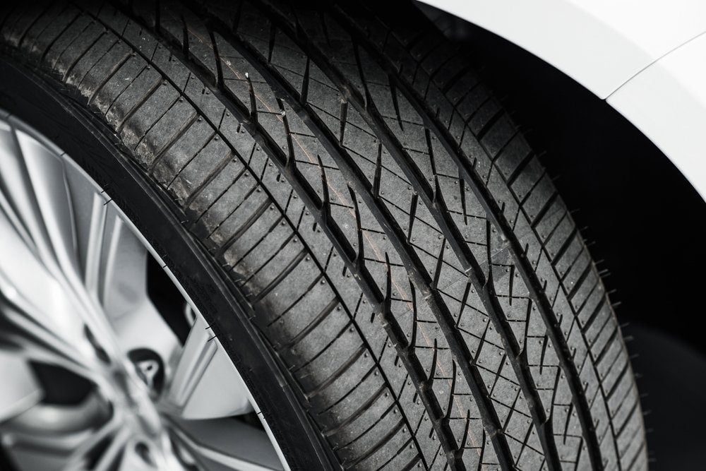 A car tire is shown, displaying detailed tread patterns. It rests against the backdrop of a vehicle's metallic rim in a well-lit environment