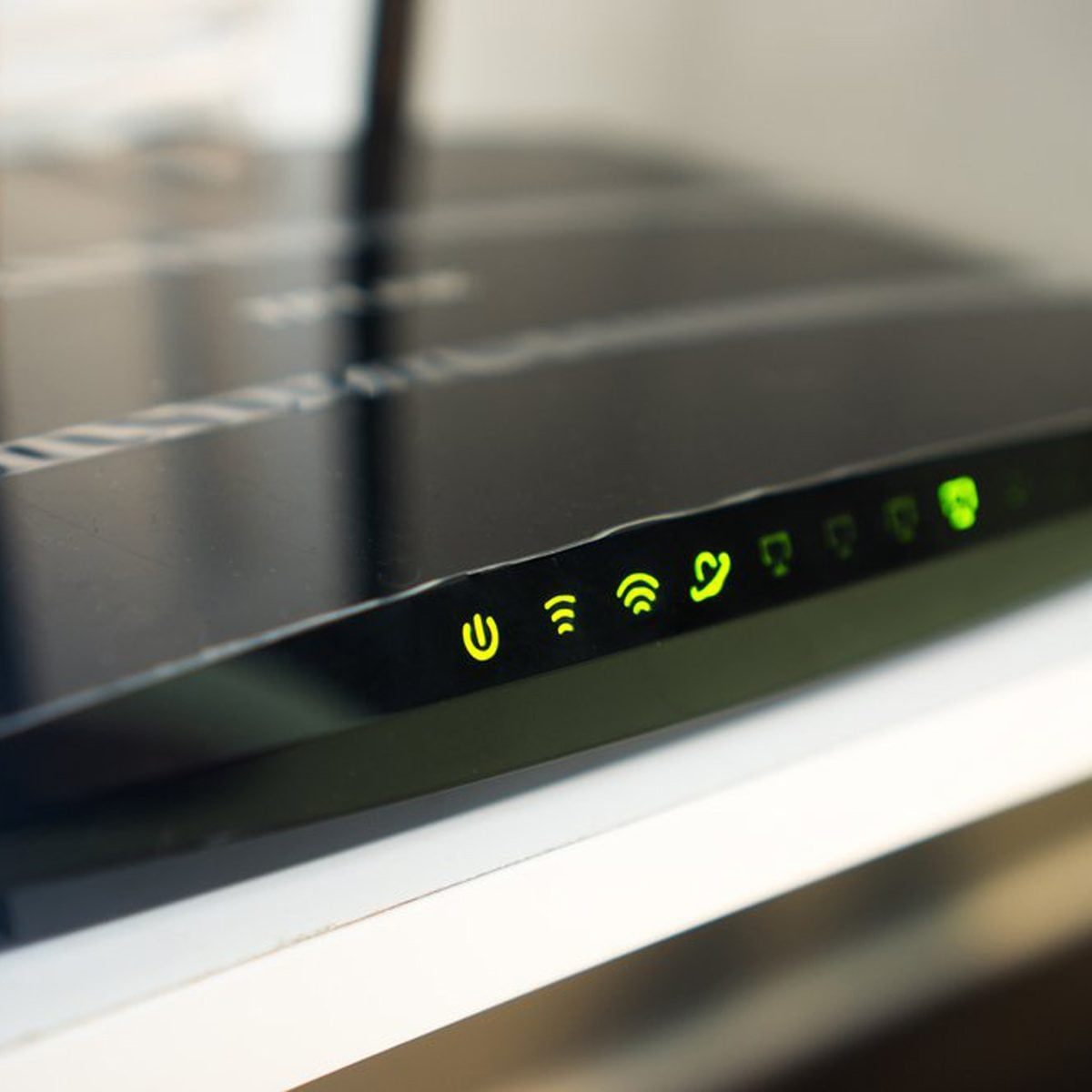 A black router sits on a shelf, illuminated by green indicator lights, signaling power, Wi-Fi, and connection status in a neutral room.