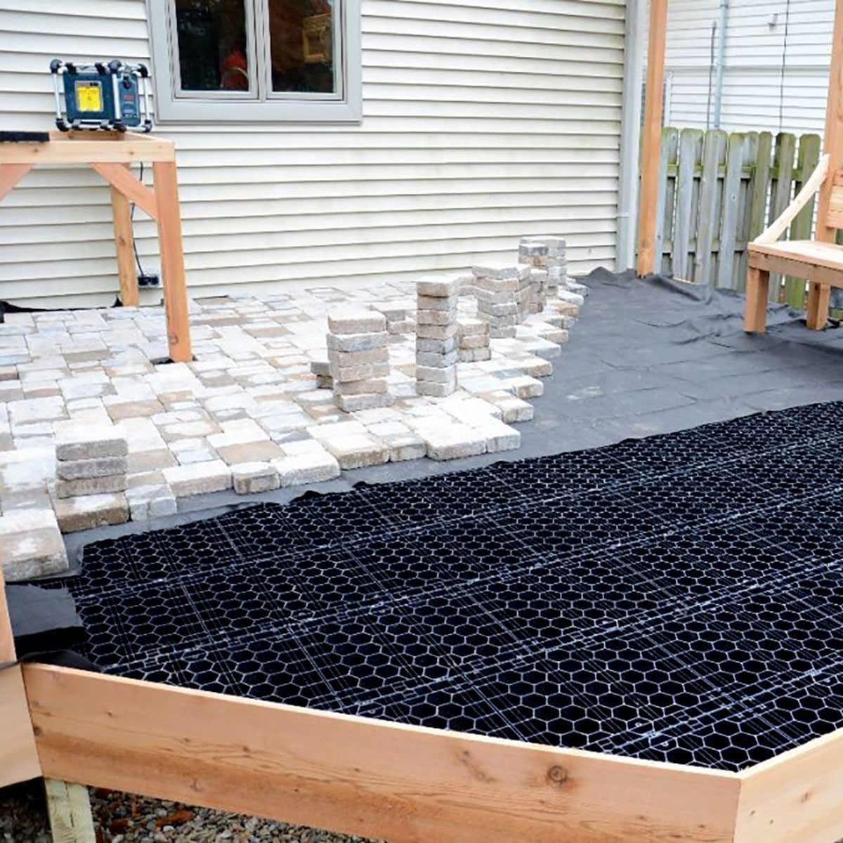 Blocks are being arranged to form a patio surface on a raised wooden deck, surrounded by landscaping fabric and construction equipment.