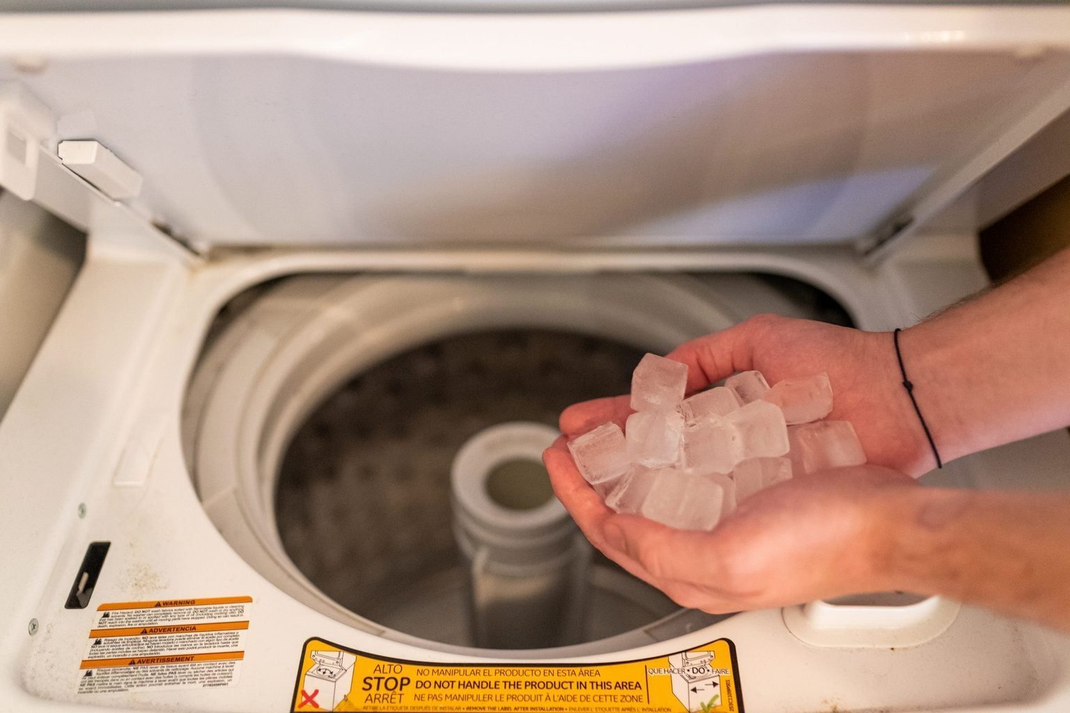 Hands hold ice cubes, placing them into a washing machine’s drum, surrounded by a warning label at the machine's edge.