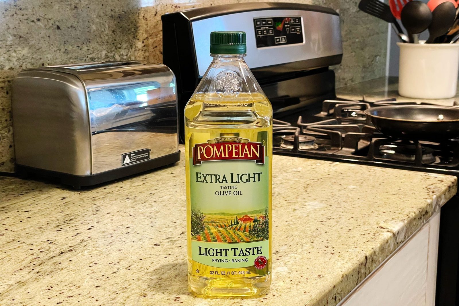 A bottle of Pompeiian extra light olive oil stands on a kitchen counter next to a toaster and a stove with a frying pan.