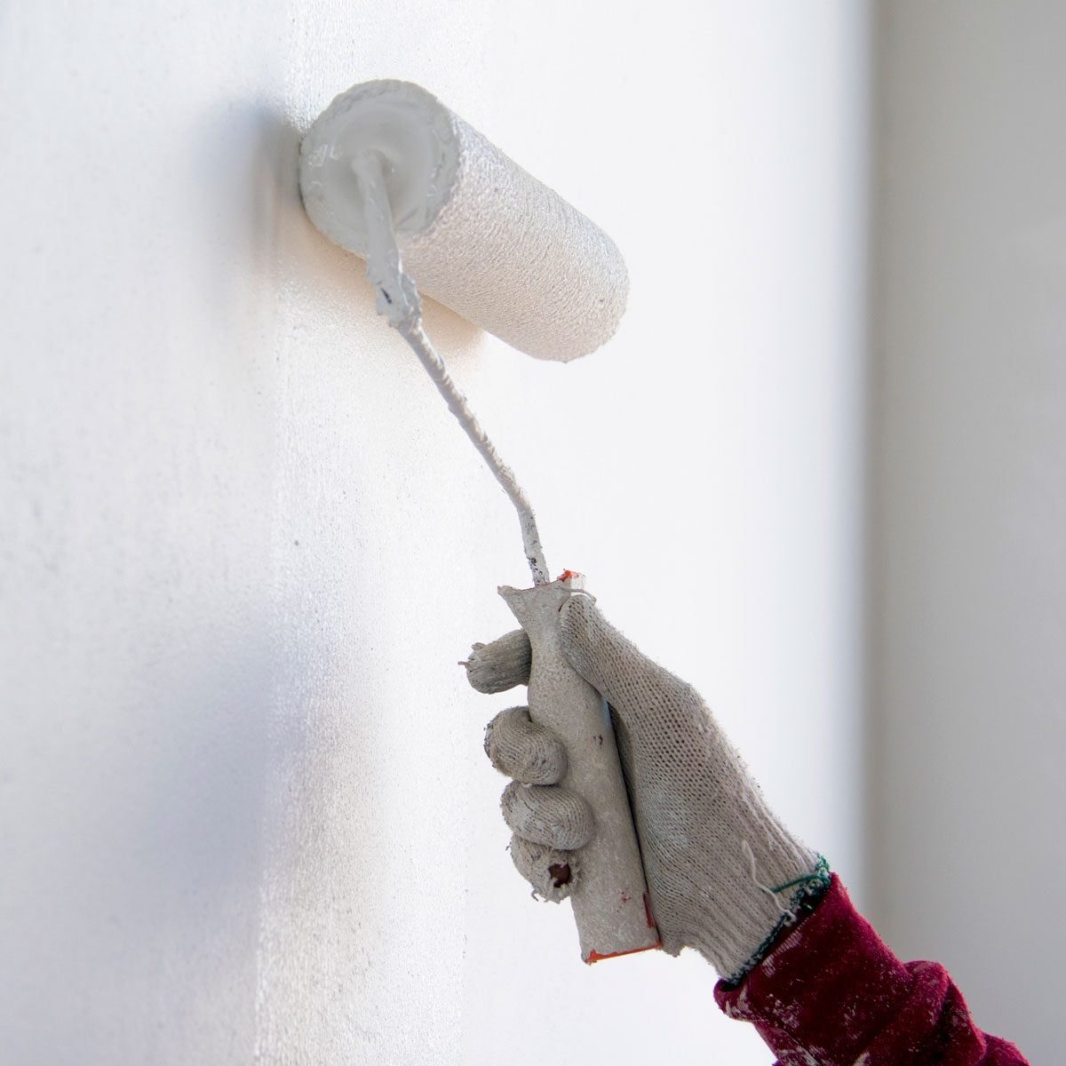 A gloved hand holds a paint roller, applying white paint to a smooth wall in a bright, indoor environment.