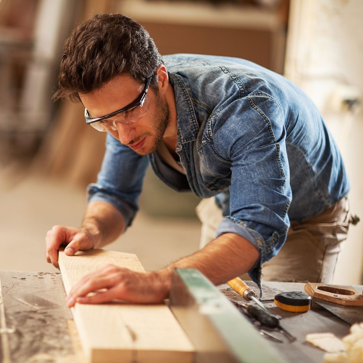 A man in safety glasses measures a wooden plank on a workbench, surrounded by tools, focused on shaping the material in a workshop environme