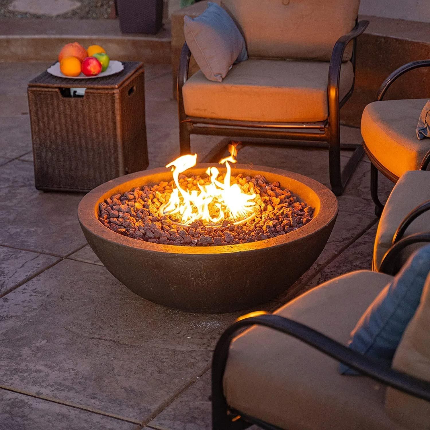 A round firepit emits flames, surrounded by smooth stones, seating nearby invites relaxation in a cozy outdoor setting. A plate of fruit res