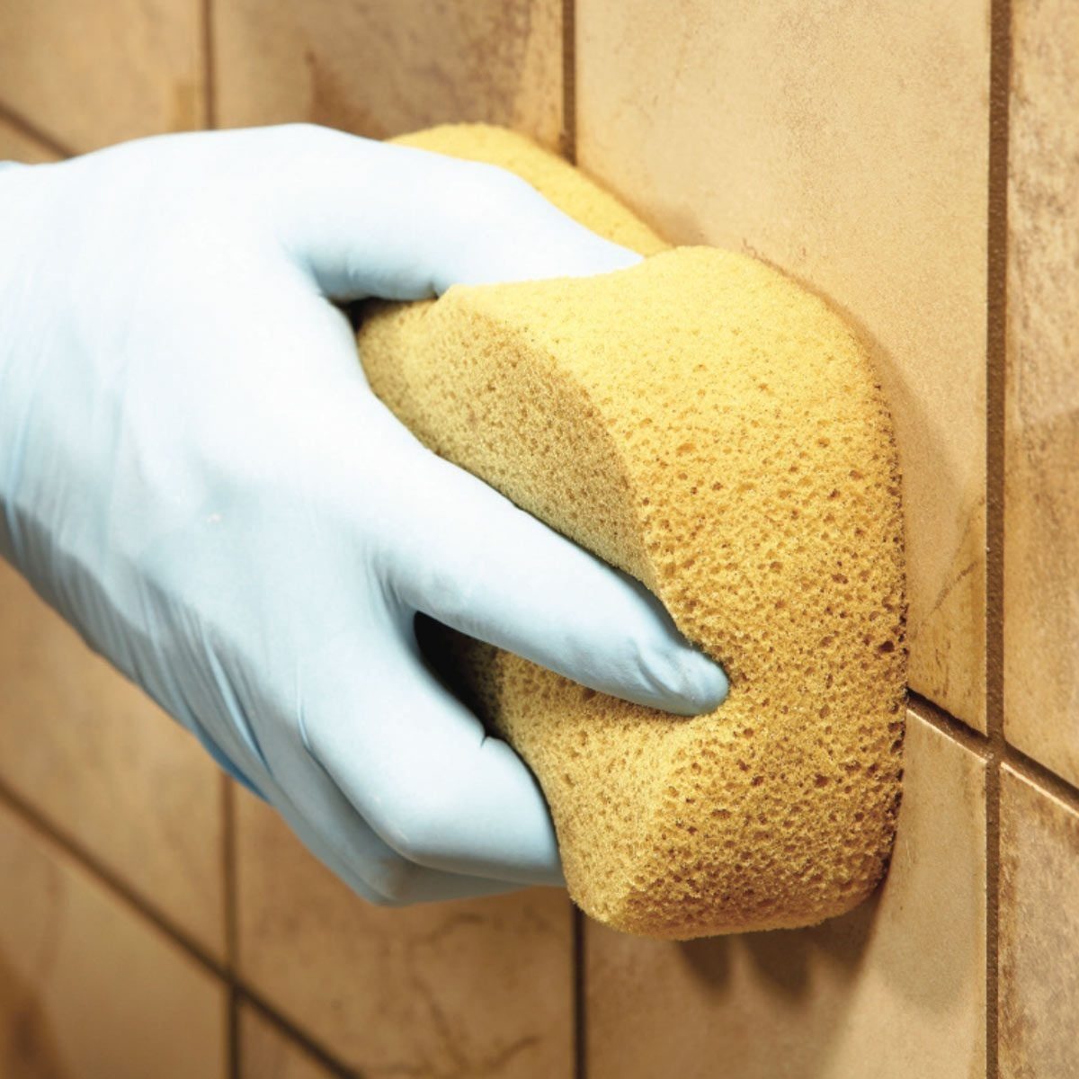 A gloved hand scrubs a sponge against a tiled wall, removing dirt or stains in a bathroom or kitchen environment.