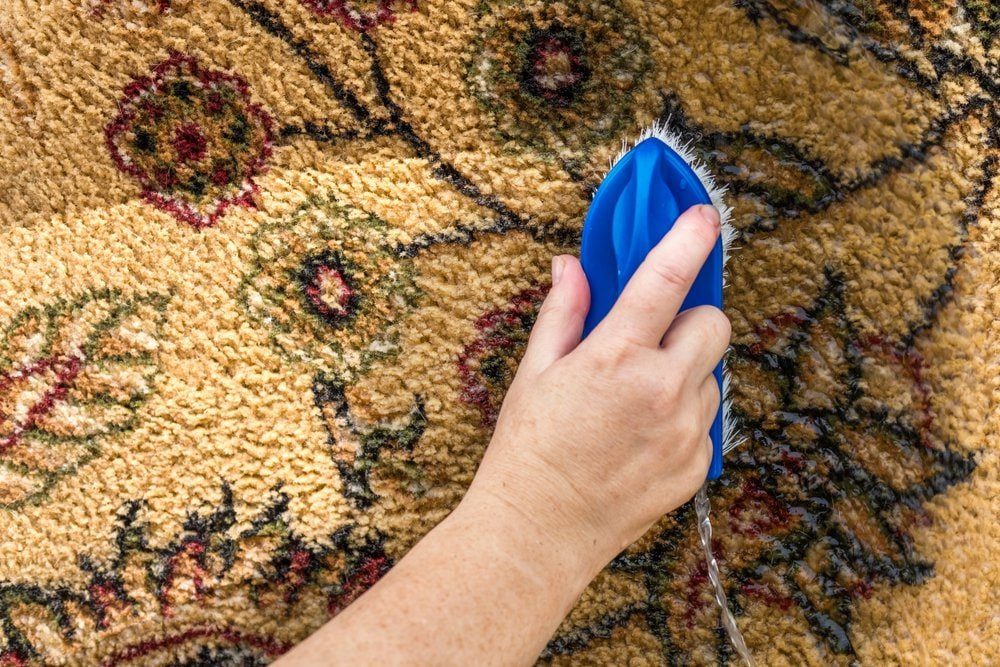 A hand holds a blue brush, scrubbing a textured yellow rug with floral patterns, focusing on cleaning the fabric.