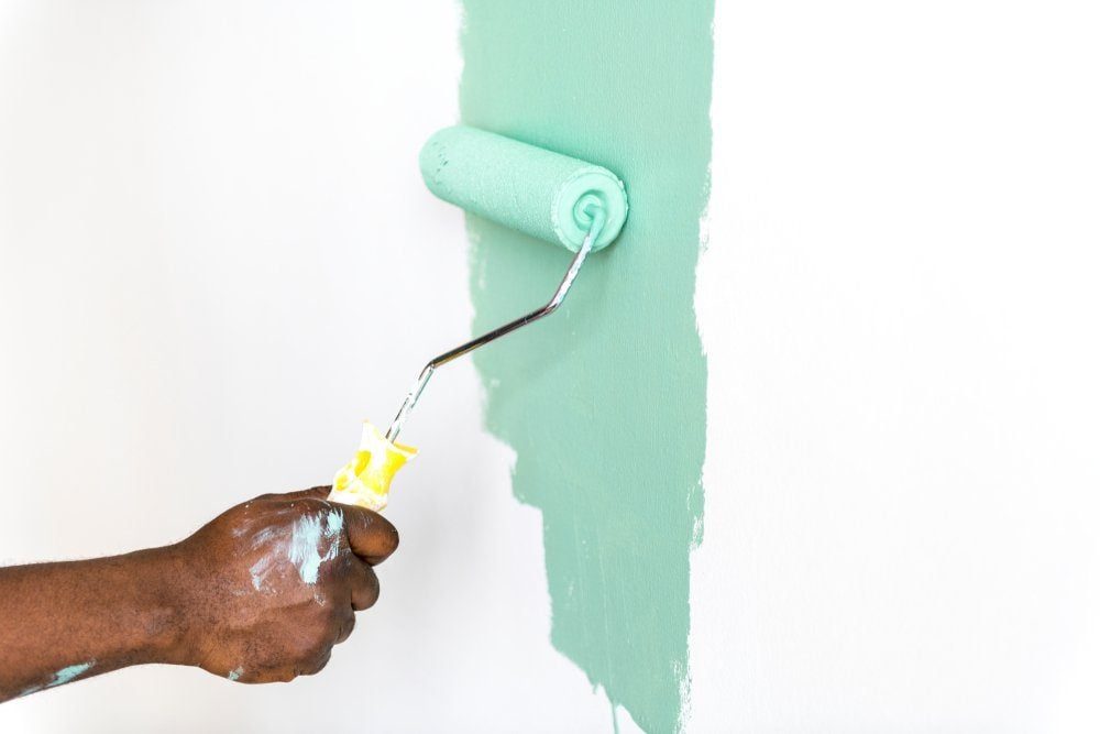 A hand holds a paint roller applying light green paint to a white wall, creating a fresh, vibrant contrast in an interior space.