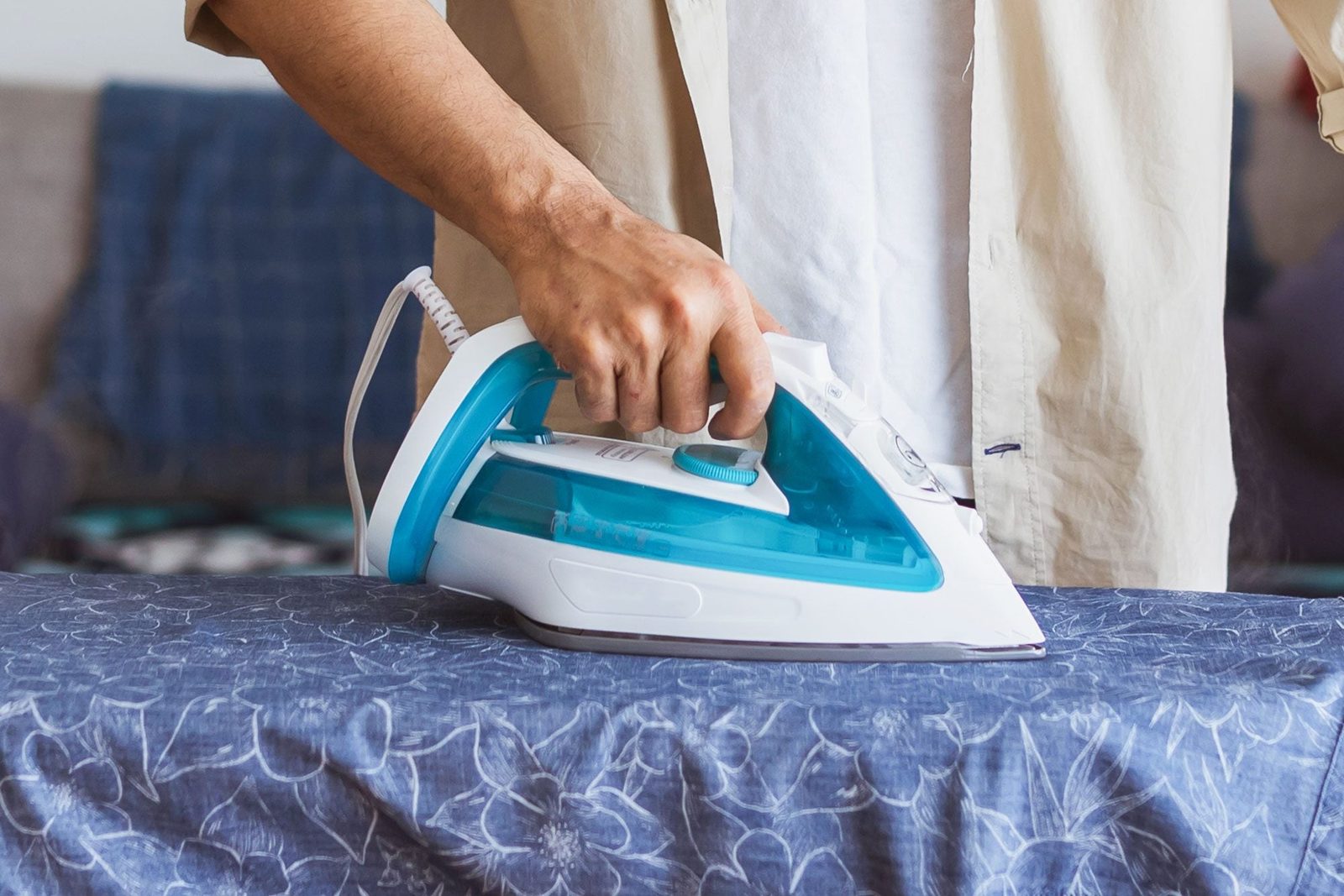 An iron glides over a patterned blue fabric on an ironing board, being held by a person in a light-colored shirt, in a casual indoor setting