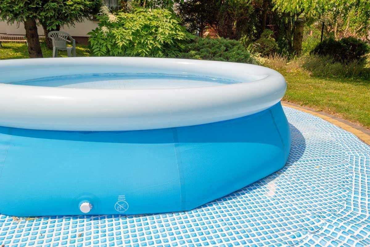 A blue inflatable pool sits on a textured surface, filled with clear water in a sunny garden surrounded by greenery and a chair nearby.