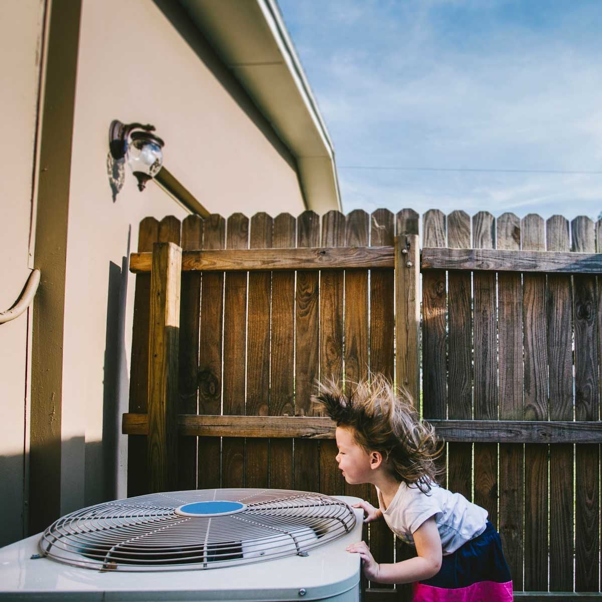 A child leans over an outdoor air conditioning unit, hair blown upward by the airflow, with a wooden fence and blue sky in the background.