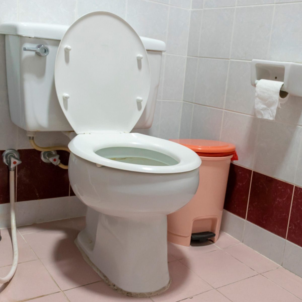 A white toilet with the lid open sits on tiled flooring, next to a small trash bin and toilet paper holder in a bathroom environment.