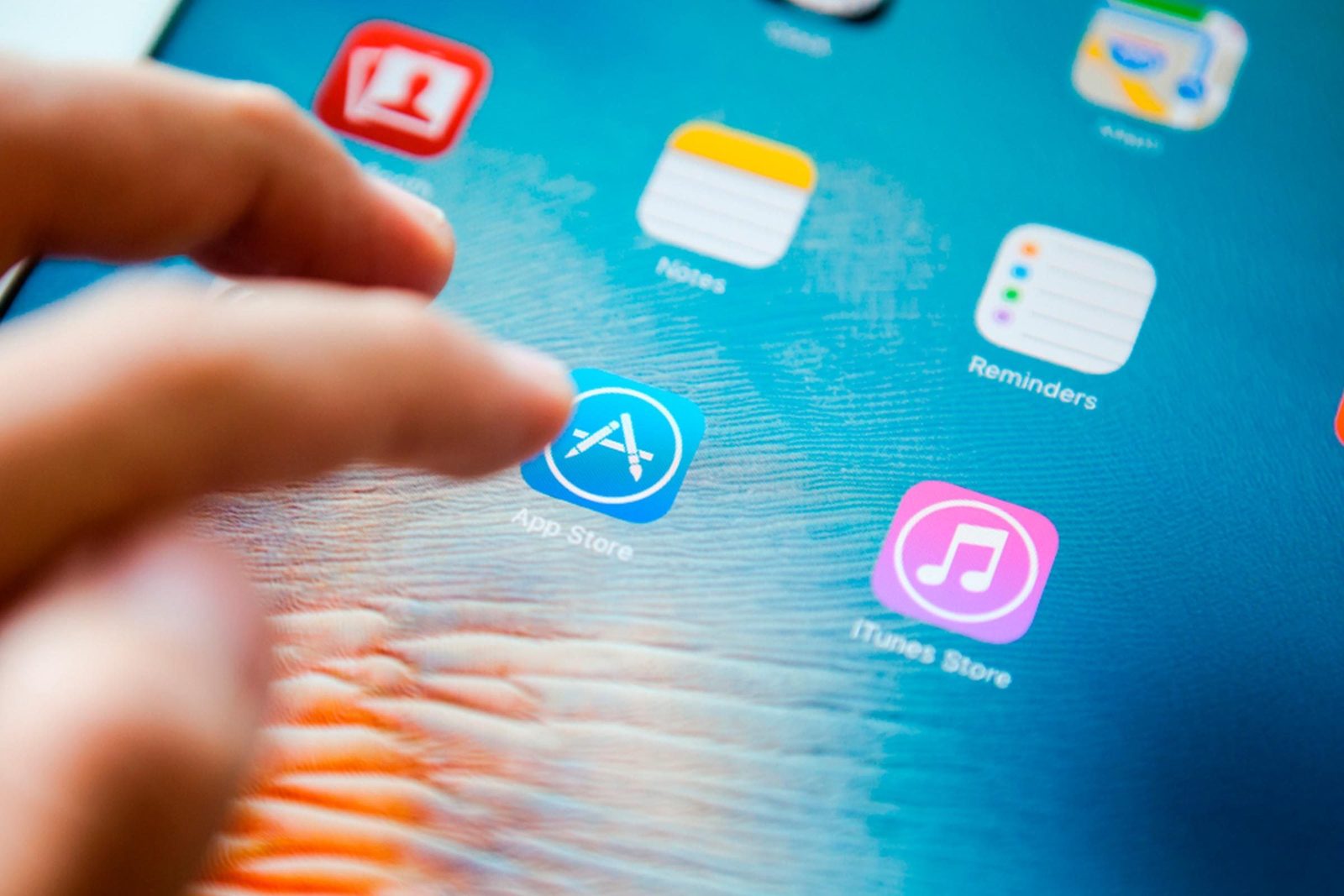 A finger taps the App Store icon on a tablet screen, surrounded by various app icons against a colorful background.