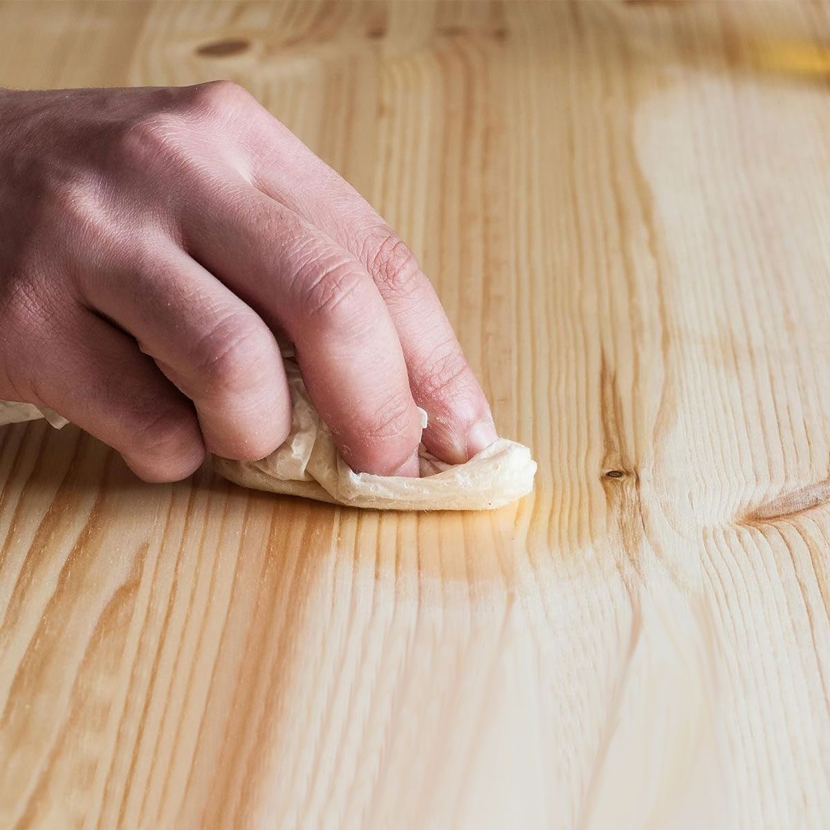 A hand wipes a cloth across a wooden surface, cleaning or polishing it, surrounded by the natural grain of the light-colored wood.