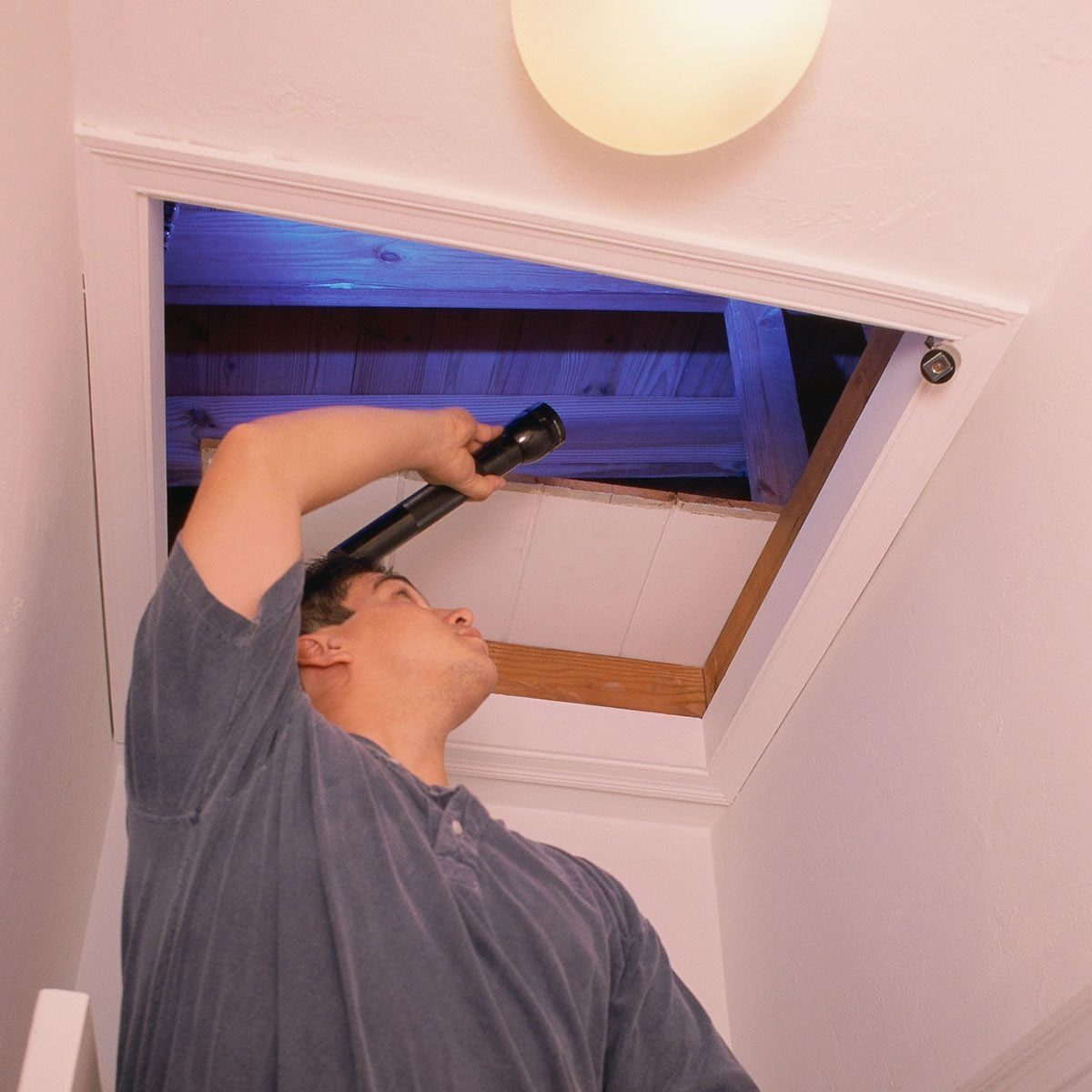 A person holding a flashlight inspects an attic opening, revealing wooden beams above, in a well-lit indoor space.