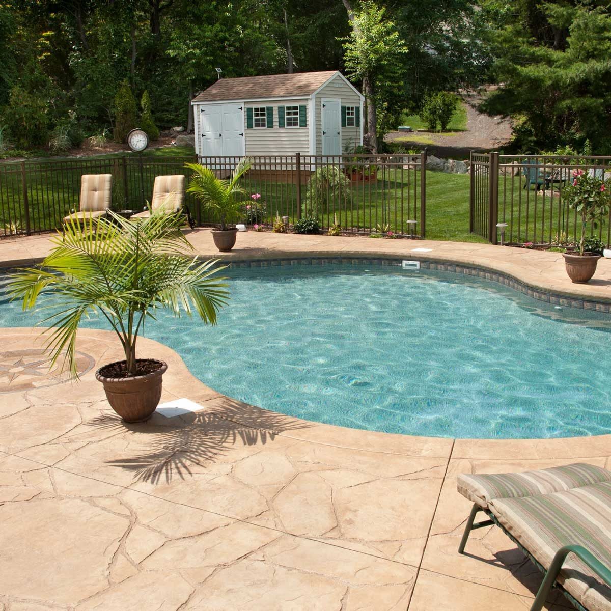A swimming pool with clear blue water is surrounded by a patio and a green lawn, featuring a small shed, potted plants, and lounge chairs.