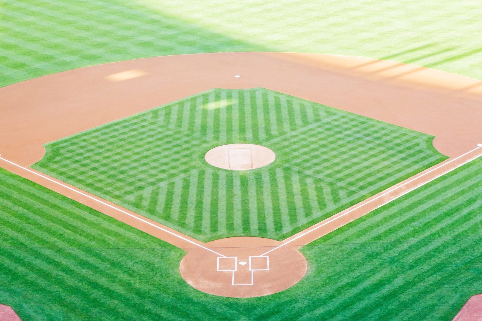 A well-maintained baseball field features a dirt infield, a pitcher's mound, and green grass with stripes, set in a sunny, open space.