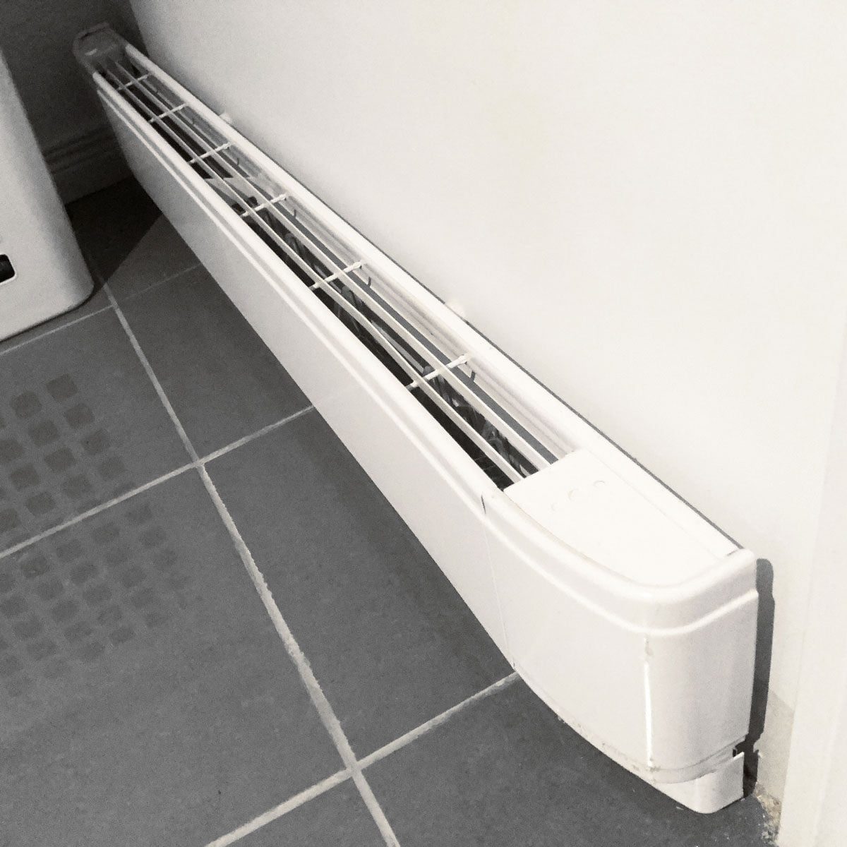 A white radiator sits along a wall, emitting warmth onto neutral tiled flooring in a modern indoor space.