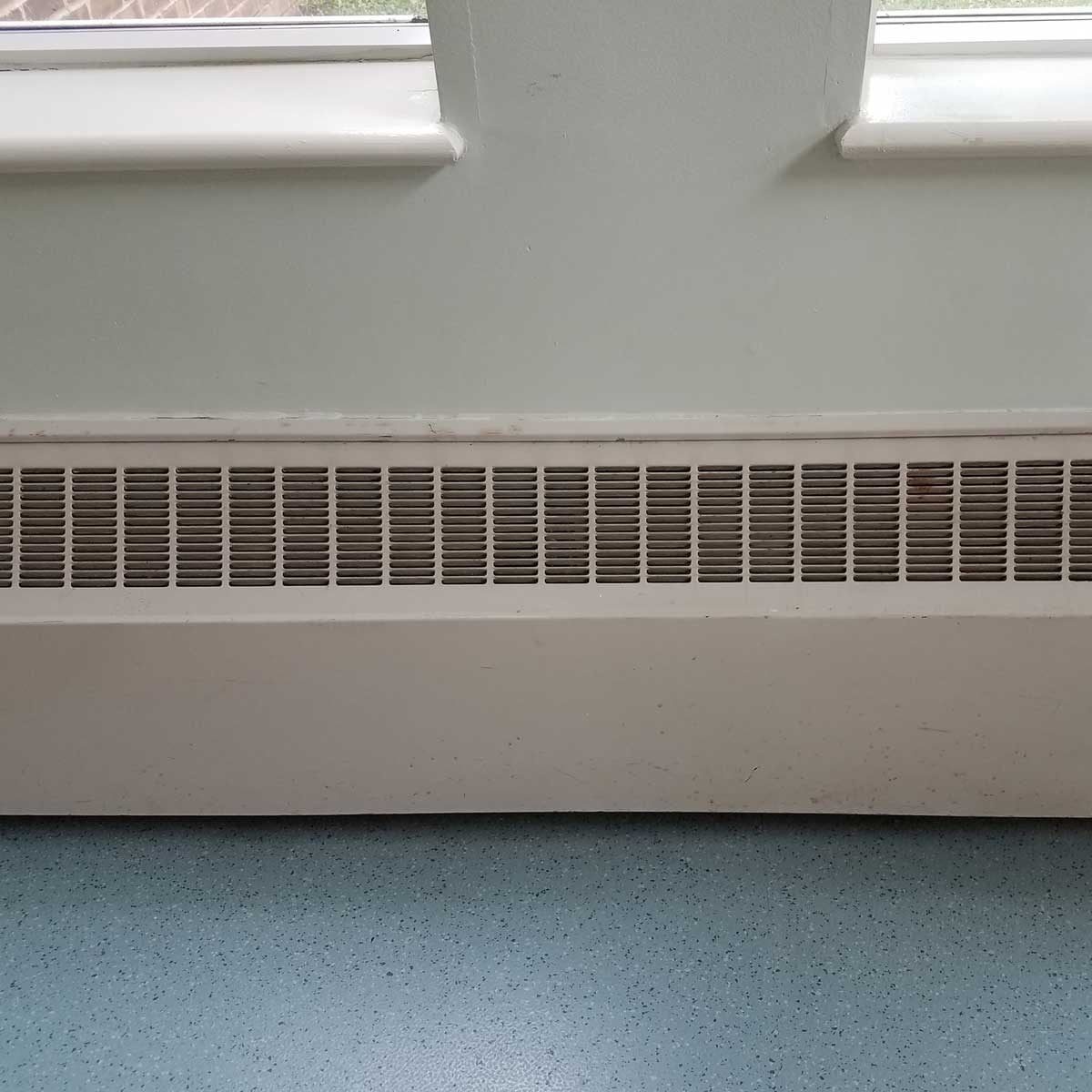 A metal heating vent is mounted along a wall, releasing air. Bright light enters through a nearby window in a room with blue flooring.