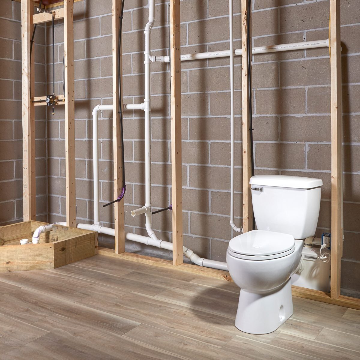 A white toilet sits on a wooden floor, surrounded by exposed plumbing and a partially finished wall of concrete blocks and wooden studs.