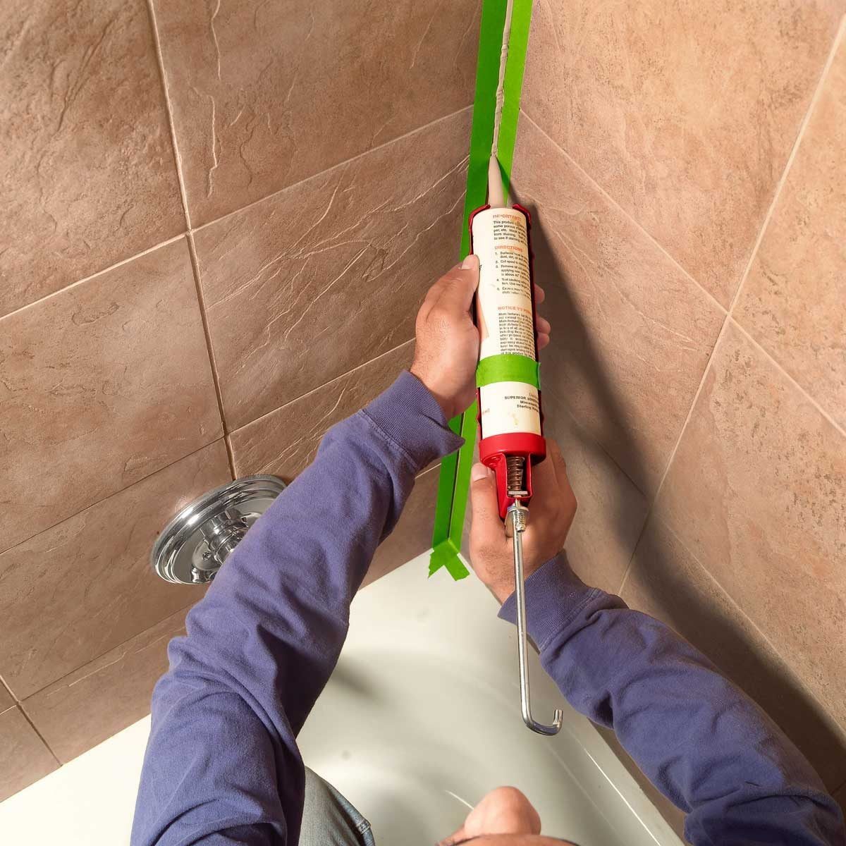A person is applying caulk from a red tube along a green tape line in a bathroom, with tiled walls and a bathtub visible.