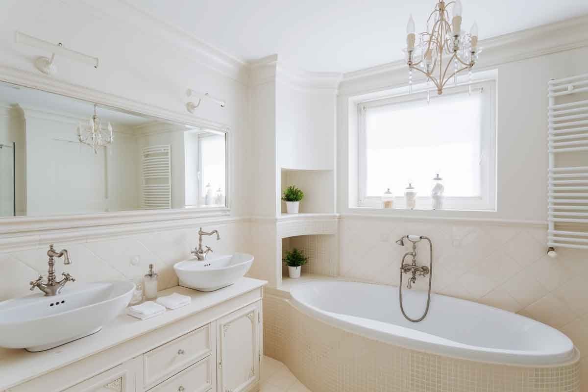 A white, elegant bathroom features a curved bathtub, two sinks with ornate faucets, and natural light from a window, enhancing the serene at