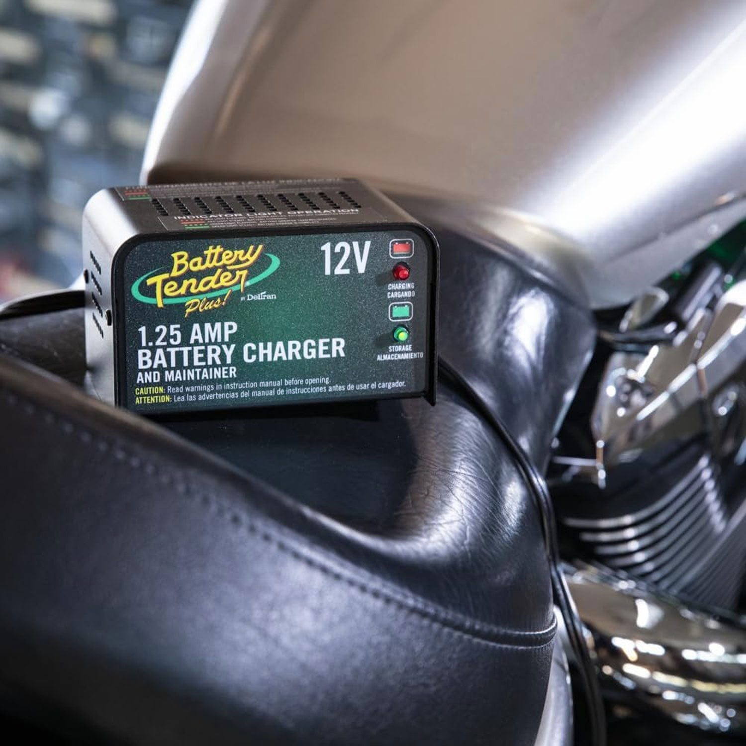 A battery charger sits on a motorcycle seat, connected with a cable, in a garage environment, indicating its functionality with charging and