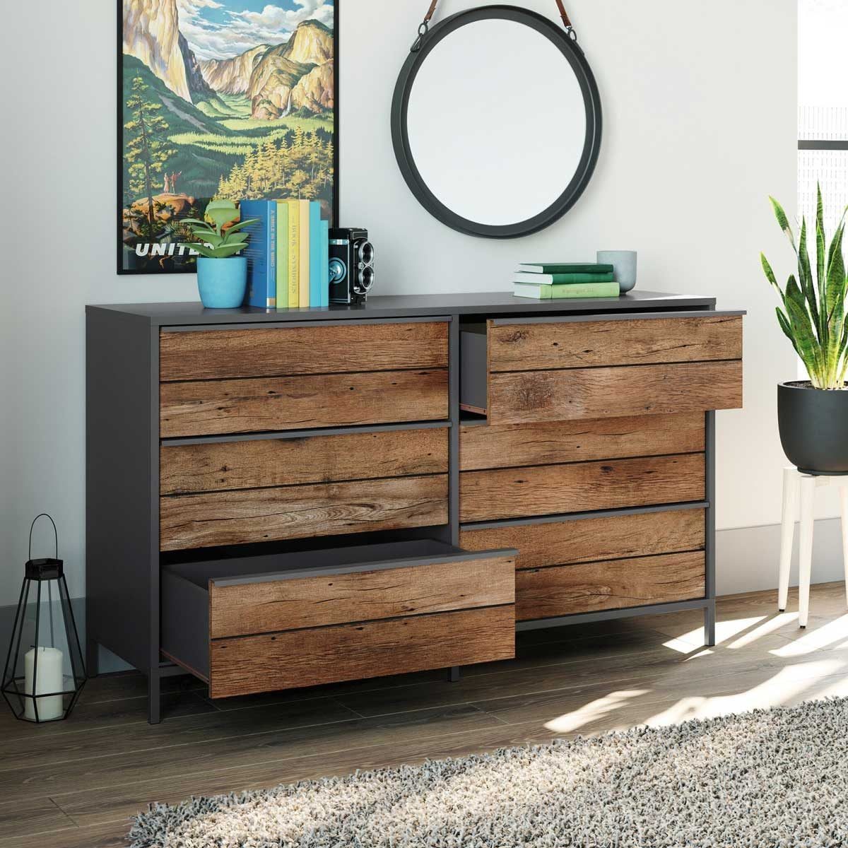 A wooden dresser with partially open drawers displays items like books and a camera, set against a wall featuring a landscape poster and a m