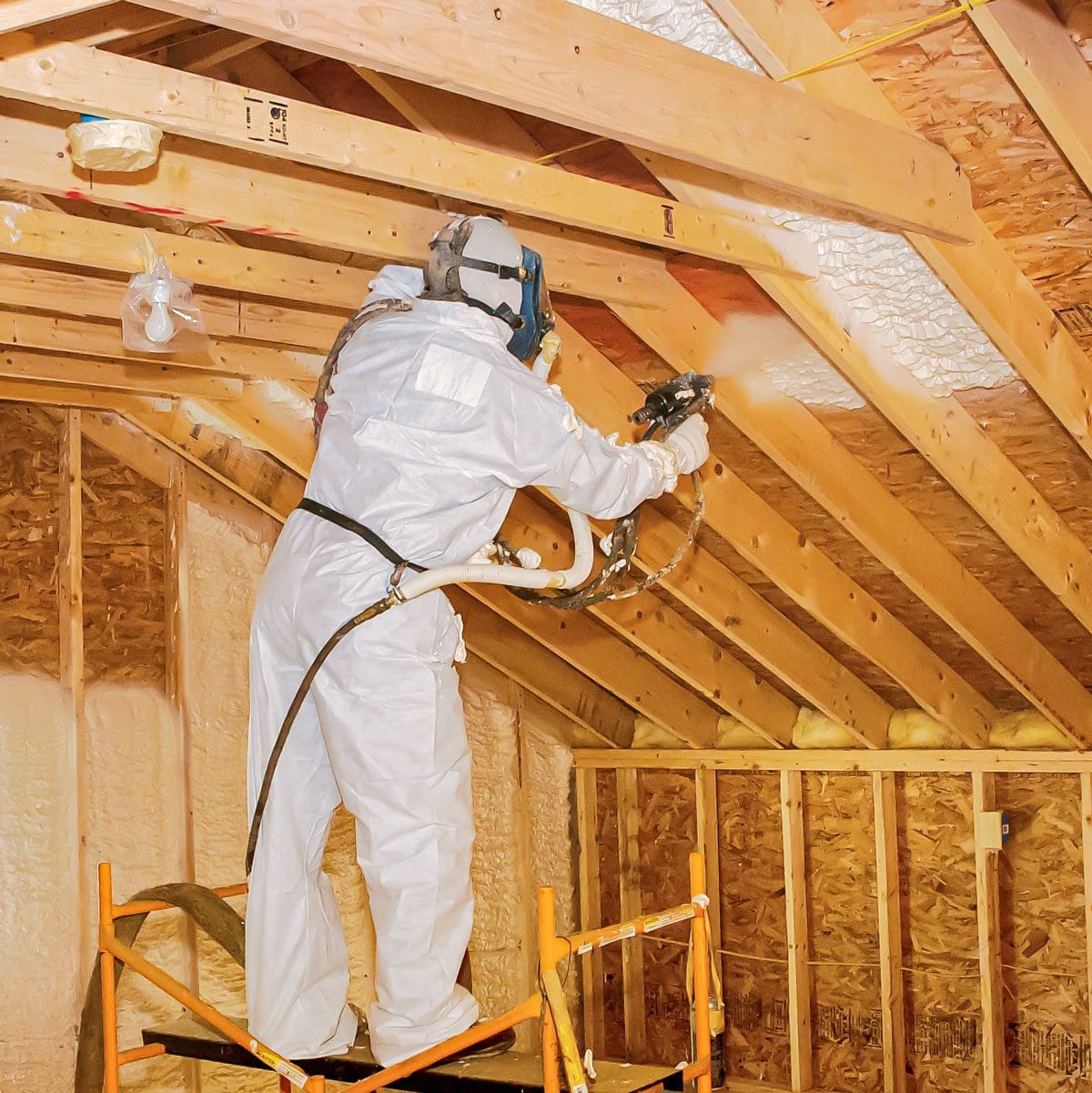 A worker in a protective suit sprays insulation on wooden rafters while standing on a scaffolding inside an unfinished attic.