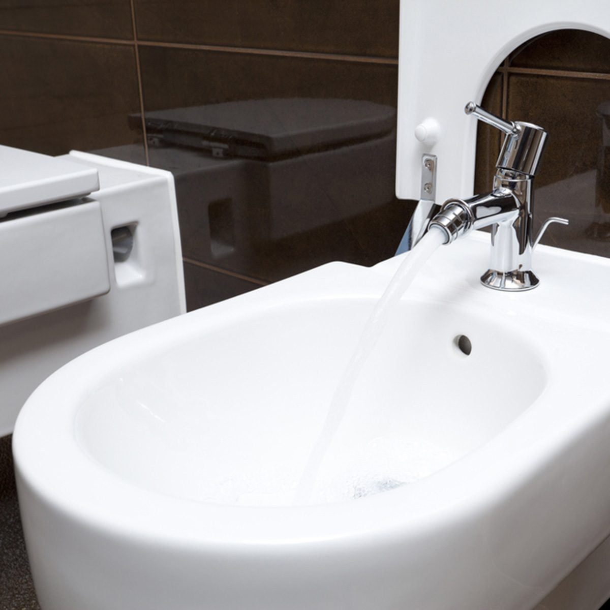 A chrome faucet releases water into a white bidet, situated in a modern bathroom with dark tile accents and a visible toilet in the backgrou