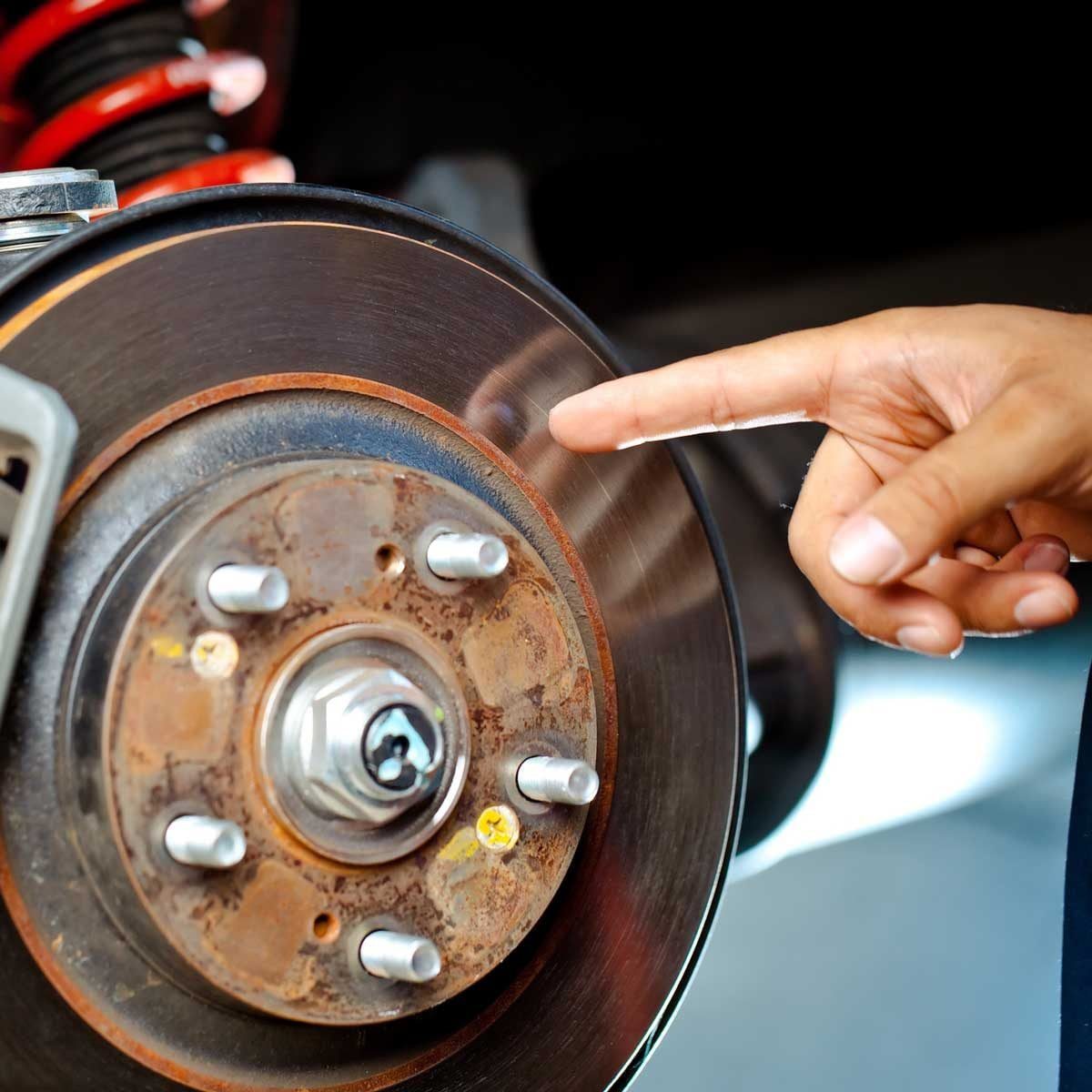 A hand touches a metal brake disc while inspecting it, situated in a well-lit garage with visible vehicle components in the background.