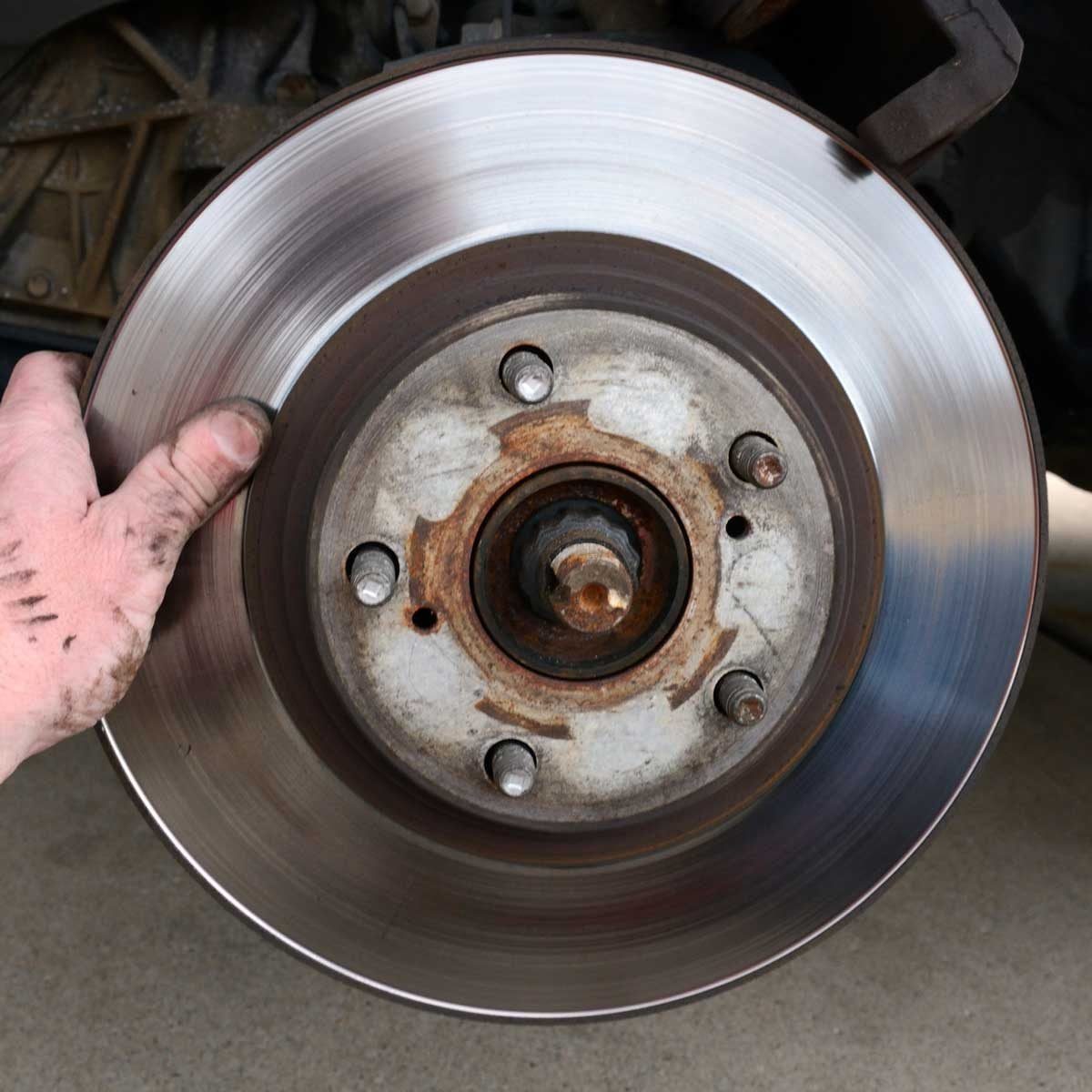 A hand holds a shiny brake rotor, showing rusted components and bolts, with a concrete surface below, indicating automotive maintenance in p