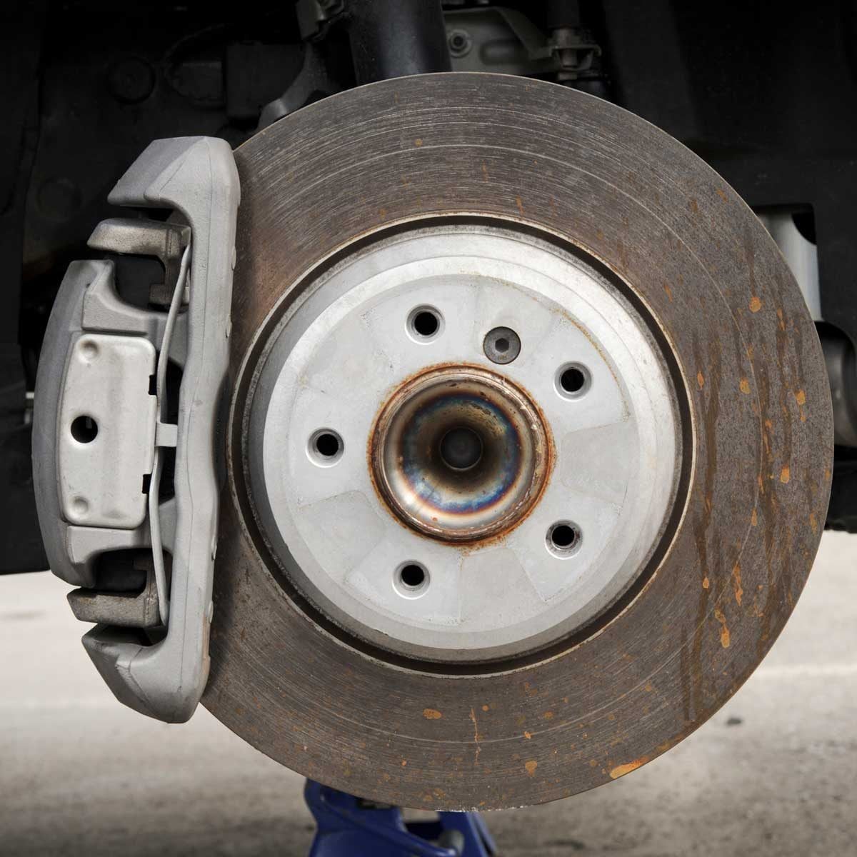 A vehicle's brake assembly is displayed, showing a disc and caliper. The brake disc shows signs of wear and corrosion, against a concrete su
