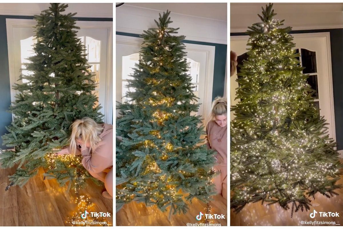 A Christmas tree is being adorned with string lights while a person crouches beside it, surrounded by a bright, warmly lit indoor space.