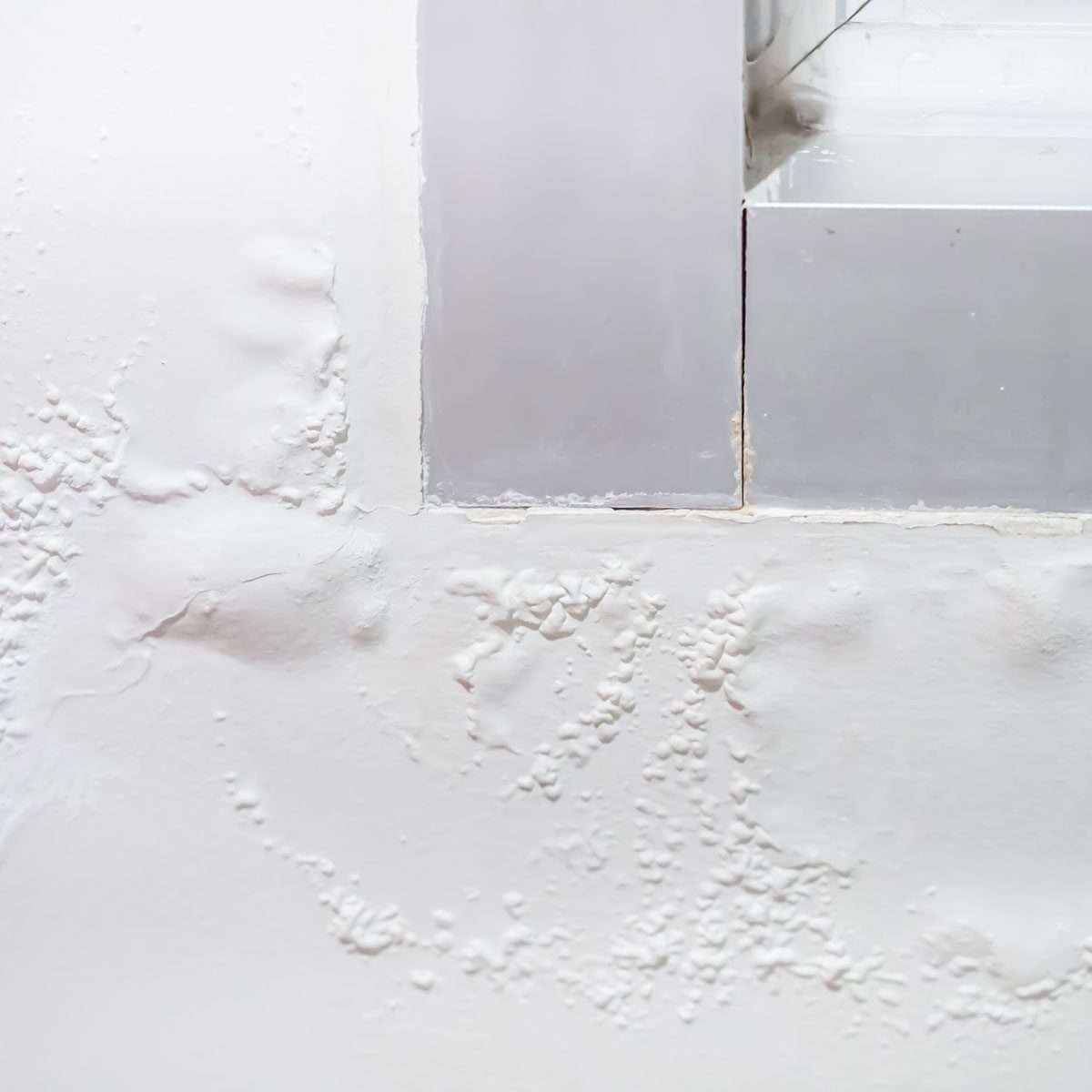 A textured white wall features a gray-framed window corner, with visible imperfections and a mix of smooth and raised surfaces creating an u