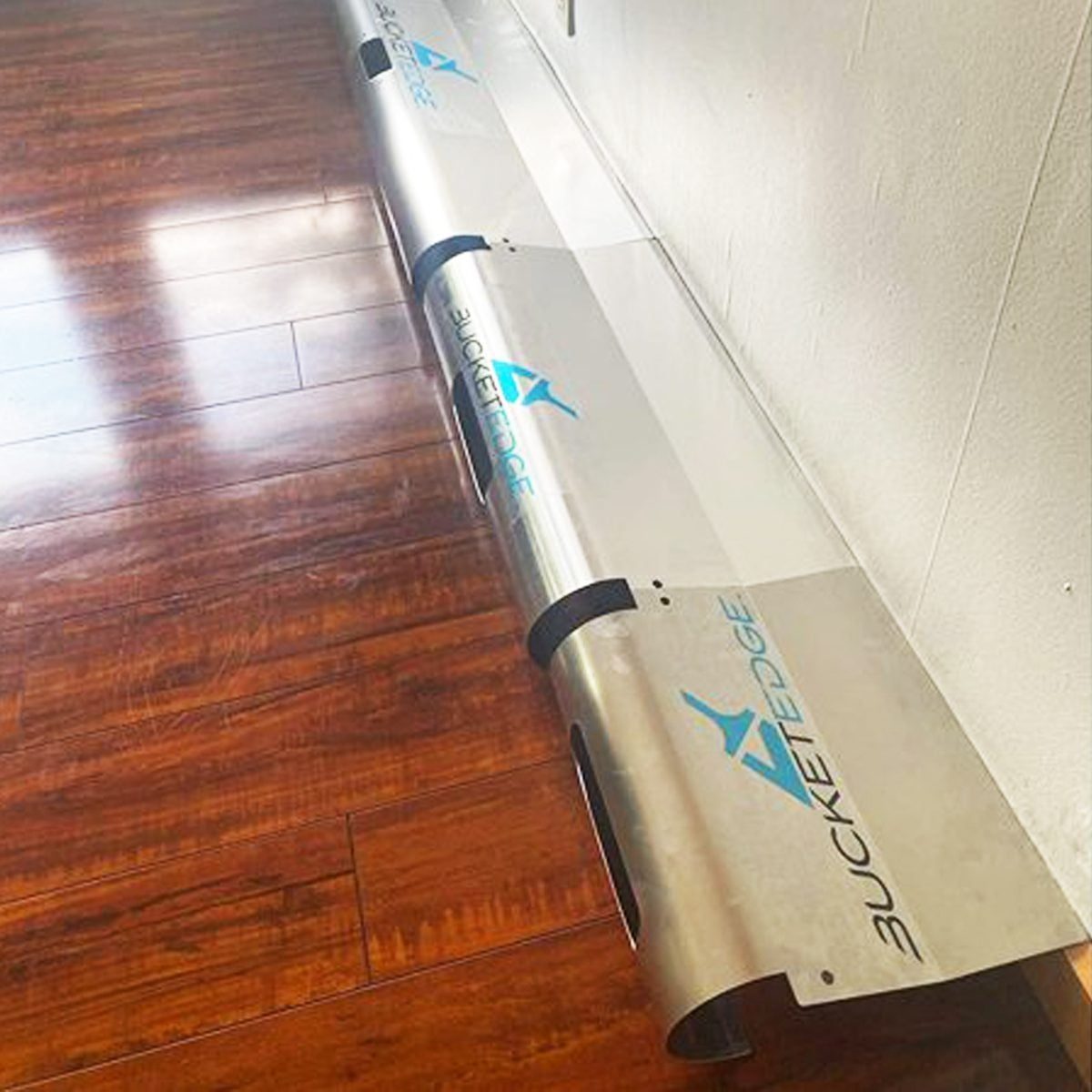 A long metal object with blue "BUCKET EDGE" branding rests against a wall on polished wooden flooring, appearing ready for installation or u