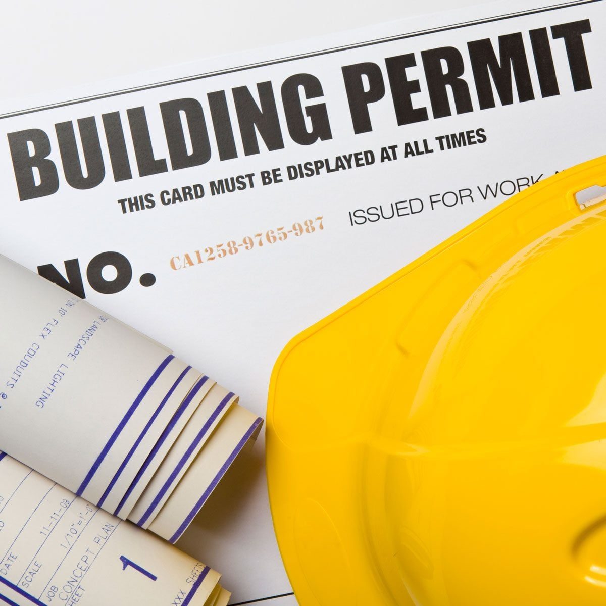 A yellow hard hat rests beside a building permit and rolled blueprint plans, indicating preparation for a construction project.