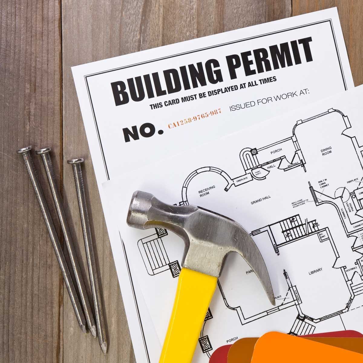 A hammer rests on a building permit and blueprint, while three nails lie beside it on a wooden surface.