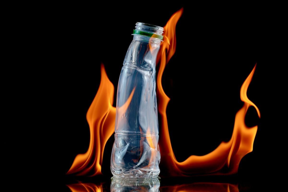 A plastic bottle stands upright while flames flicker around it, set against a dark background, creating a striking contrast.