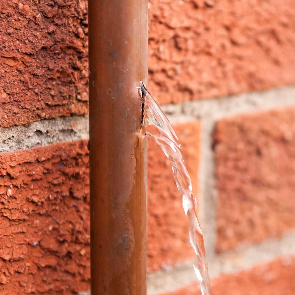 A copper pipe leaks water from a crack, flowing outwards onto a textured red brick wall behind it.