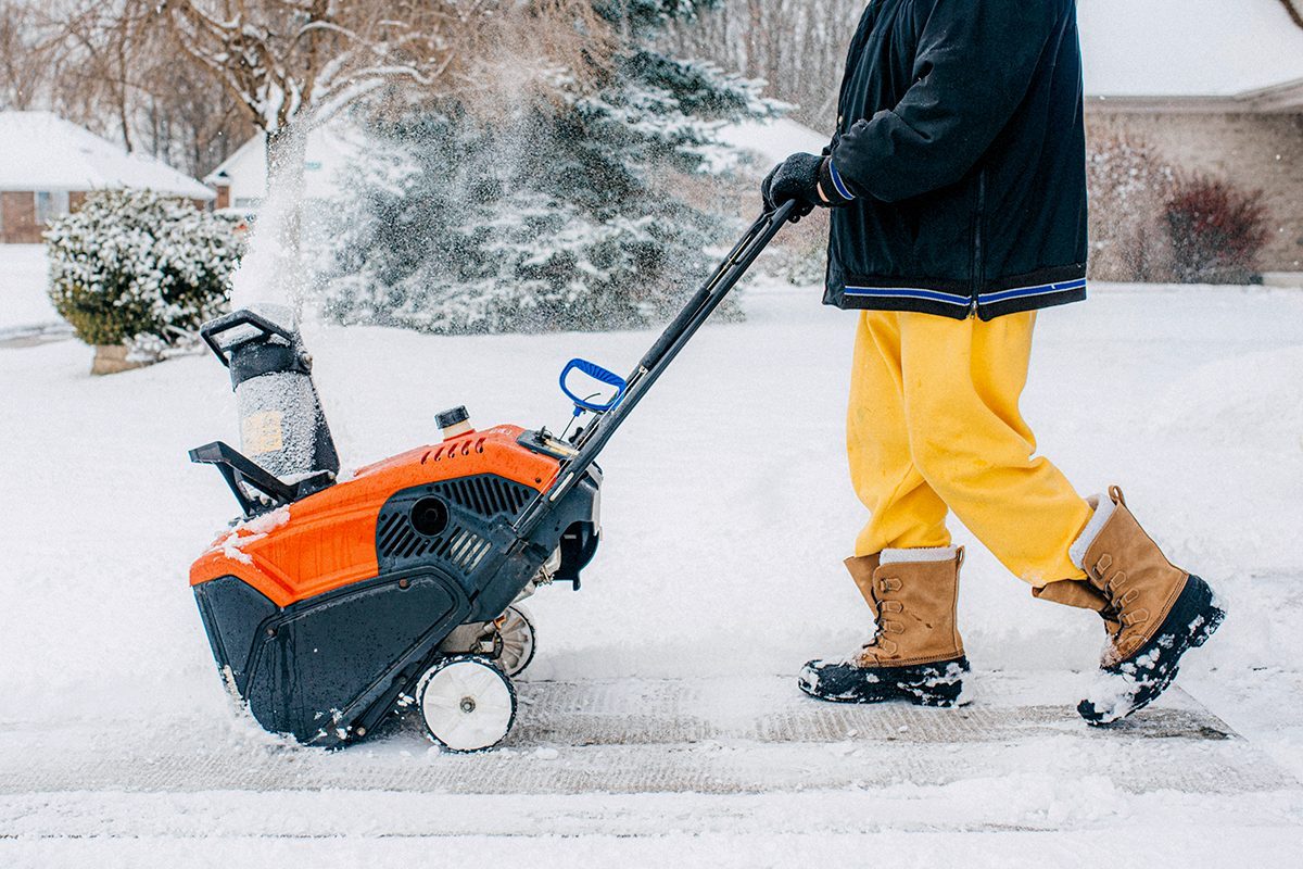 A person wearing yellow pants and brown boots uses an orange snow blower to clear snow from a sidewalk in a snowy residential area.