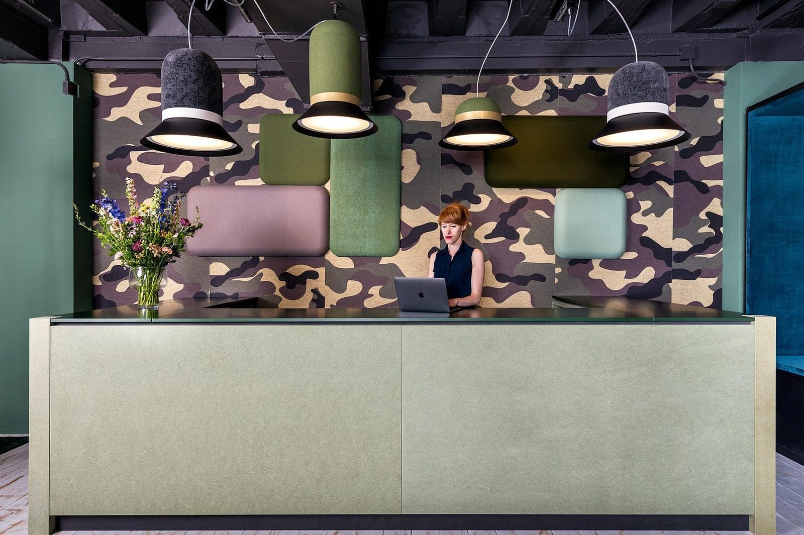 A woman sits at a sleek green reception desk, using a laptop in a modern space with camouflaged wall decor and stylish hanging lamps above.