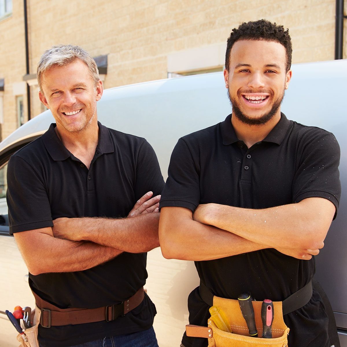 Two smiling men in black polo shirts stand with arms crossed, wearing tool belts, in front of a van and a light-colored building.