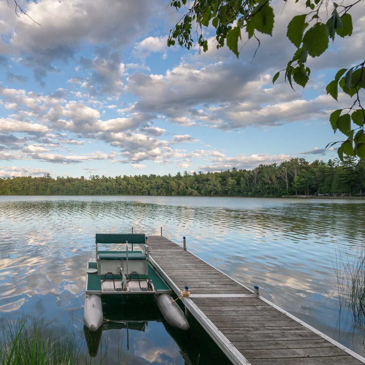 A small boat rests on a dock, reflecting in calm water surrounded by lush trees and a blue sky filled with clouds.