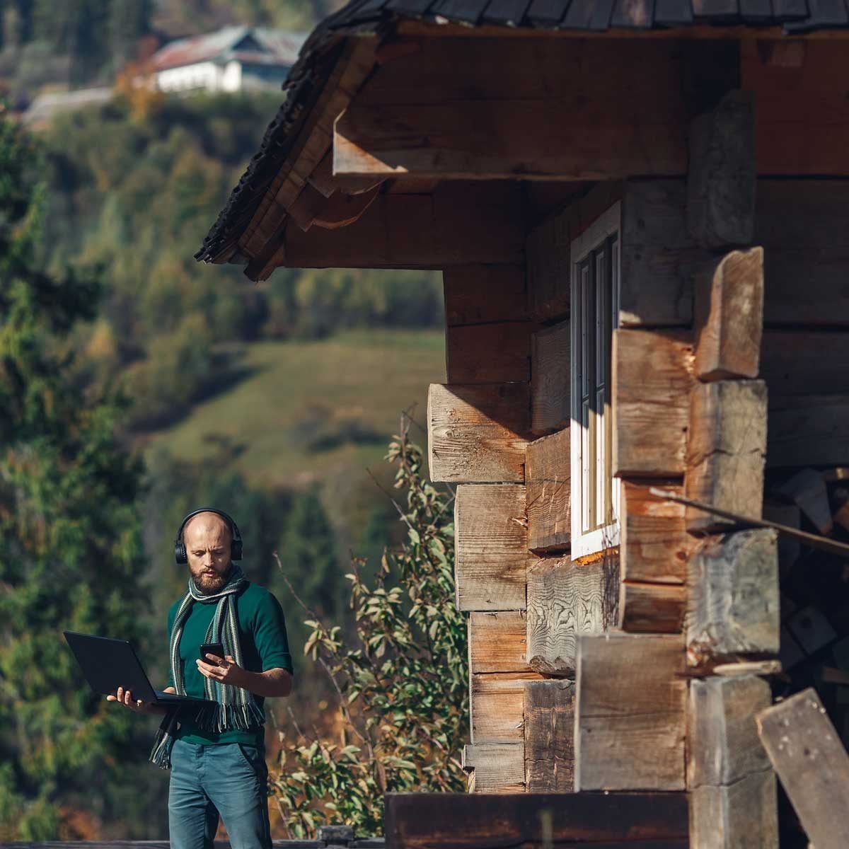 A man with headphones stands outside a wooden hut, holding a laptop and a phone, surrounded by greenery and a hilly backdrop.